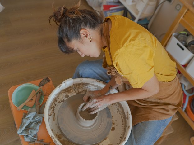 A woman uses a potting urn to shape a brown bowl in a workshop. She wears a brown apron.