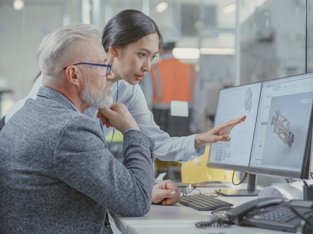 Two professionals observe a mechanical design at a work desk. They are in a workshop.