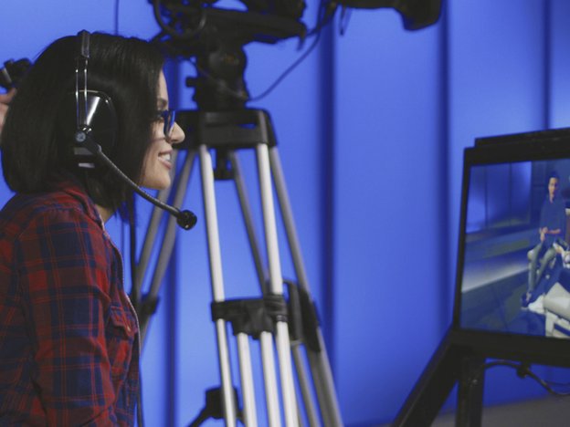 A woman watches a show being filmed. She sits beside a standing camera and wears a headset.