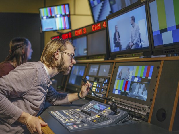 A man leans in and speak to a microphone attached to directing equipment. There is a woman beside him, a tv playing a show and they are in a studio.