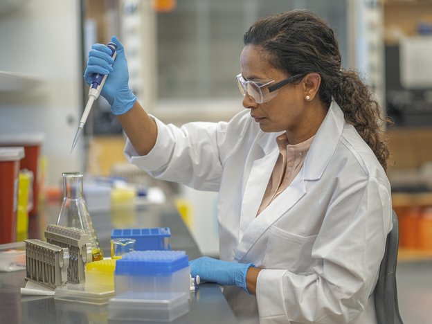 A woman puts a pipette into a beaker. She wears a white lab coat, blue gloves and safety glasses.