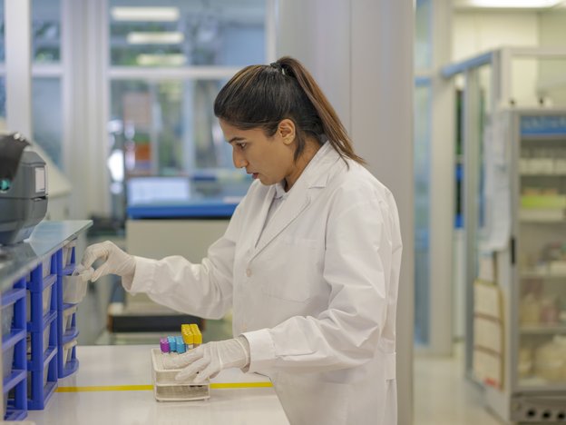 A woman takes equipment from a lab draw. She has a test tube tray and wears a white lab coat.