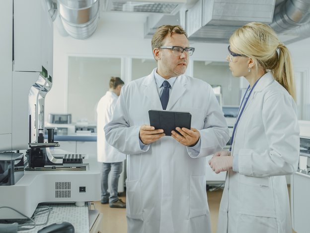 Two scientist discuss in a lab. They are dressed in white lab coats, wearing safety glasses, one is holding a tablet.