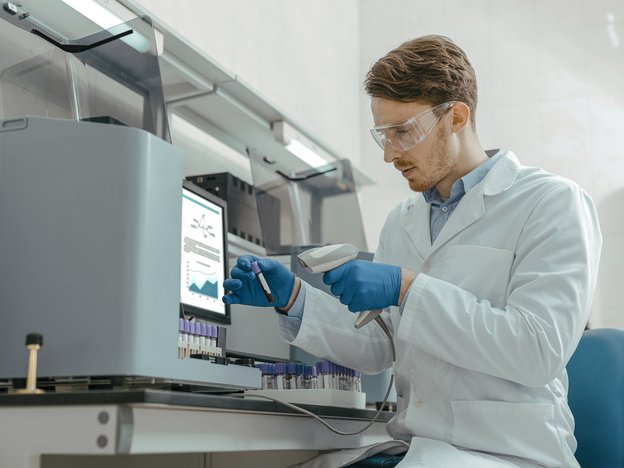 A scientist scans a beaker sample. They are sitting down wearing a white lab coat and safety glasses.