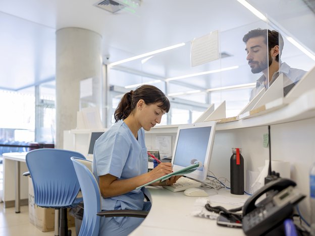 A woman writes on a clipboard. She wears blue scrubs and sits at a reception desk with a glass barrier. There is a man behind the barrier.