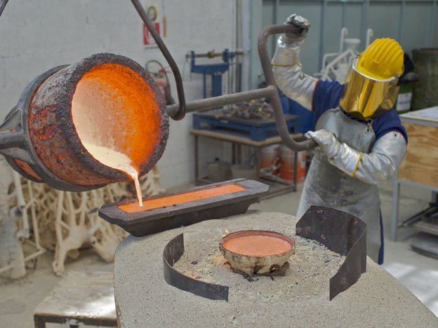 A metal caster operates a machine to pour hot metal into a tin. They wear silver protective wear, a yellow face mask and helmet and silver gloves.