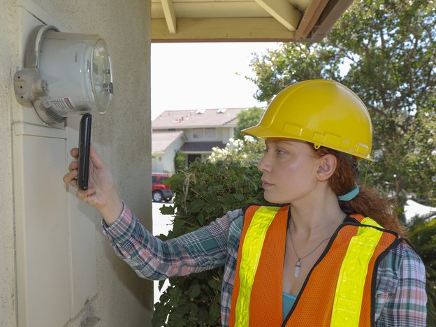 A woman checks a meter. She is outside a house. She wears an orange hi-vis vest and a yellow safety helmet.