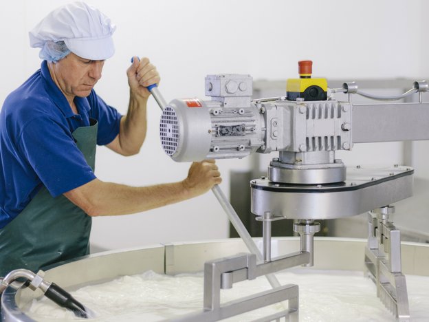 Man in a blue shirt with a green apron and white hat mixes cheese using a large mixing machine.