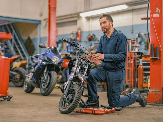 A male mechanic in blue overalls kneels on the ground to clean a motorcycle using a rag. He is in a motorcycle factory.
