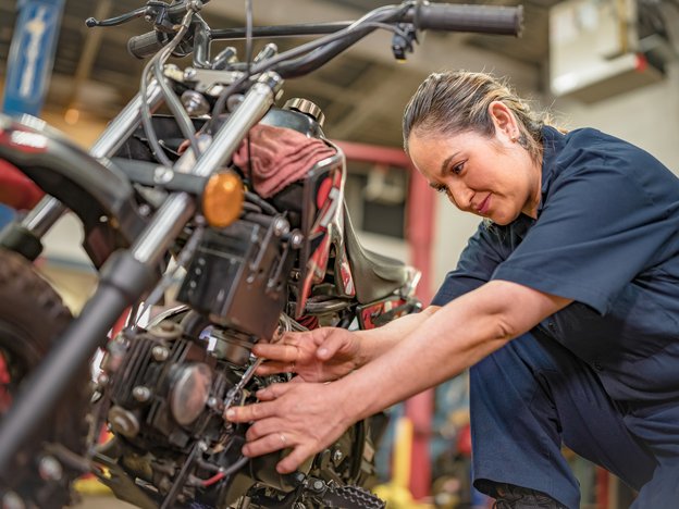 A female mechanic repairs a motorcycle in a factory. She wears blue overalls.