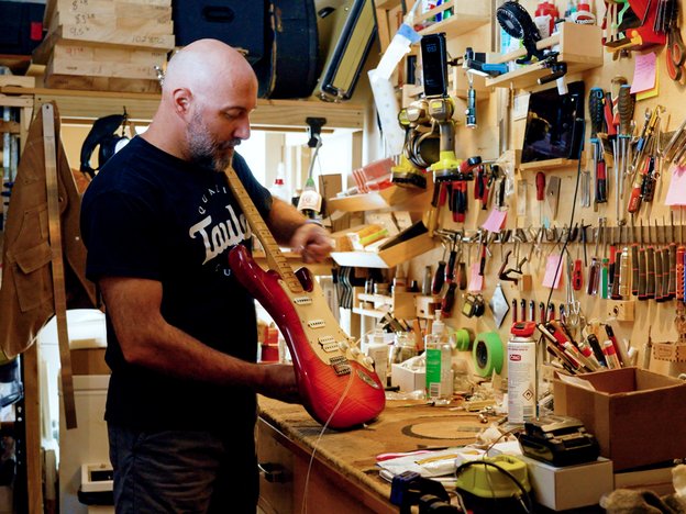A person fixes a guitar on their workbench. On the wall is a variety of tools and equipment.