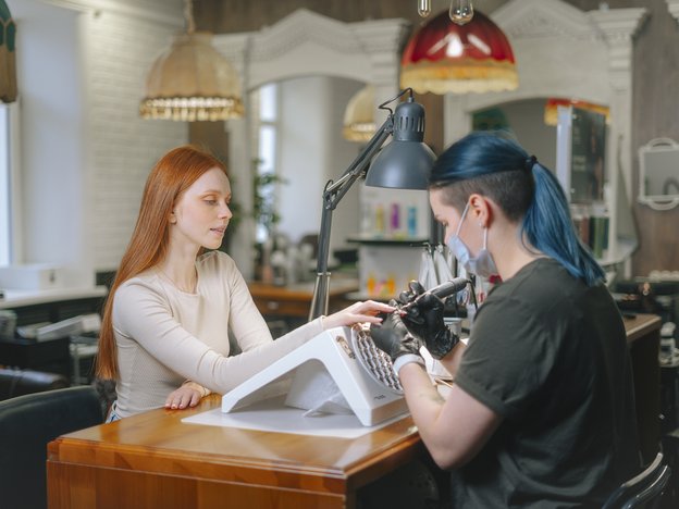 A woman gets her nails painted at a nail salon. The nail technician is using a nail brush and is wearing gloves and a facemask. They are sitting opposite each other at a desk in a nail salon.