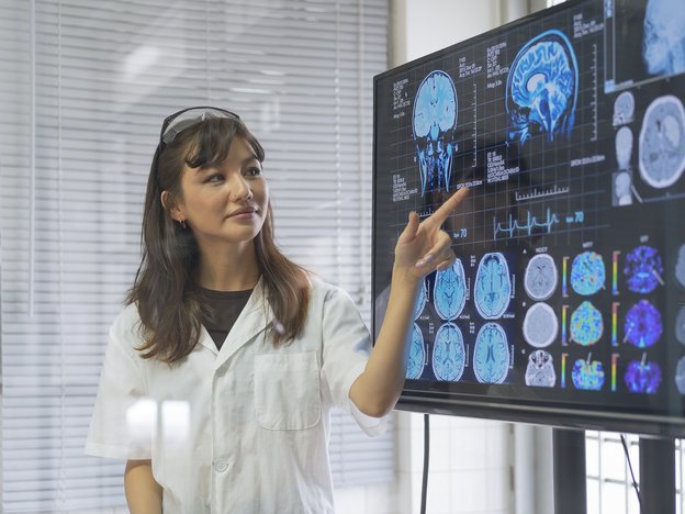 A female neurologist points to computer scans of the brain on a monitor screen. She is wearing a white shirt in a medical room.