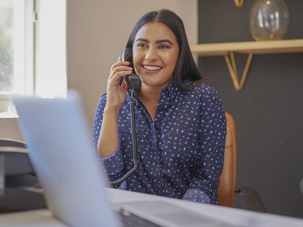 A woman holds a landline phone to her ear. She sits at an office desk and there is an open laptop on the desk