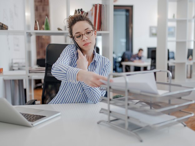 A woman sits at a desk in an open plan office. She holds a mobile phone to her ear and places paper onto a paper tray. There is an open laptop on the desk.
