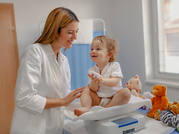 A female pediatrician checks out a baby in a medical room. She wears a white doctor's coat. The baby sits smiling, upright on some weighing scales and has a small bandage on their finger.