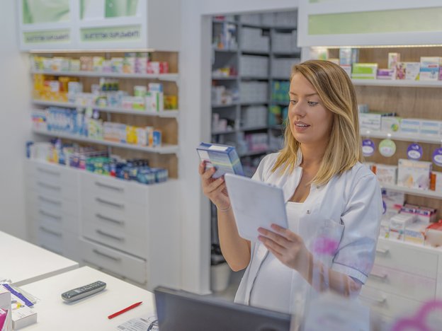A female pharmacy technician wearing a white coat looks at a box of medication in a pharmacy.