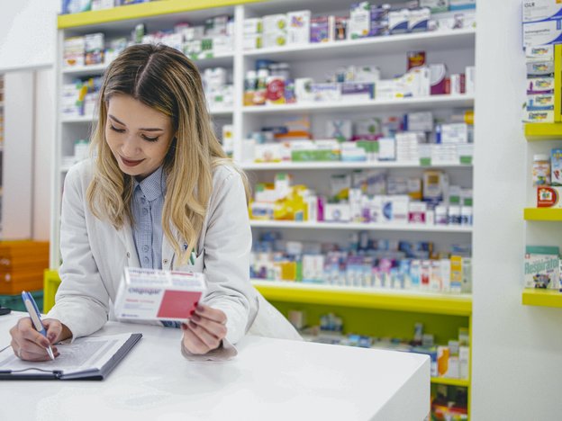 A female pharmacy technician in a white coat looks at a box of medication and take notes at a desk in a pharmacy.