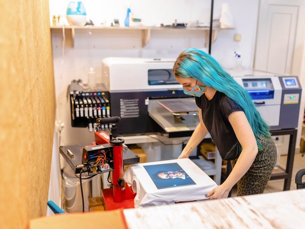 A woman adjusts an image on a t-shirt in a printing studio. She has blue hair and a blue facemask.