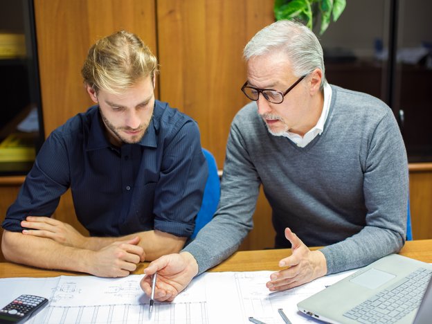Senior businessperson teaches younger trainee in a modern office. They are sitting at a desk looking at plans.