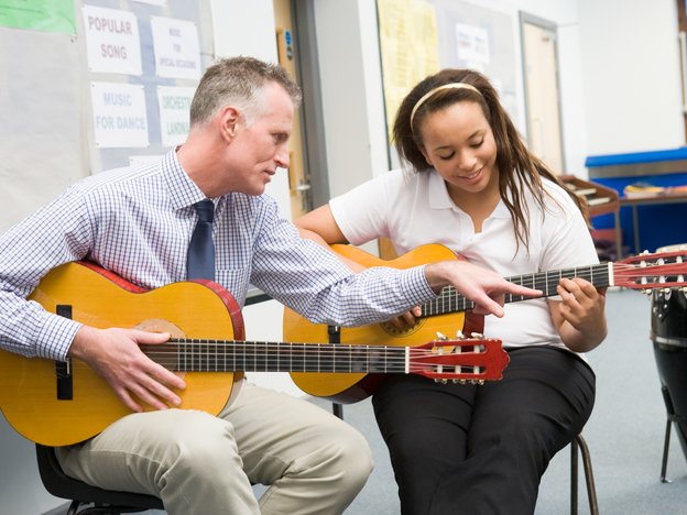 A man teaches a girl how to play guitar. They both hold a guitar of their own.