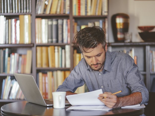 A man reads a document at his office desk. There is an open laptop and a mug on the desk.