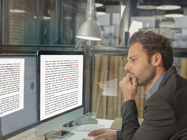 A man reads a document on a computer monitor. He sits at a desk.