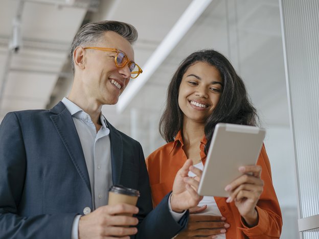 A woman holds a tablet with one hand and stands next to a man who holds a takeaway coffee cup in one hand. They look at the tablet.