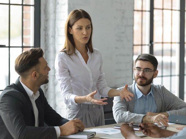 A woman stands and gestures to a group of people sat near a table. She stands in between two men and there is a third but can only see their hands.