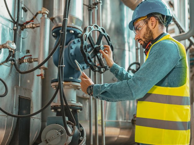 A man operates a pump in a factory. He is wearing a fluorescent yellow jacket, blue helmet and safety glasses.