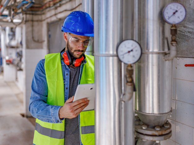 A man looks at a tablet as he inspects a pump in a factory. He is wearing a fluorescent high visibility vest, blue helmet, safety glasses and orange earmuffs.
