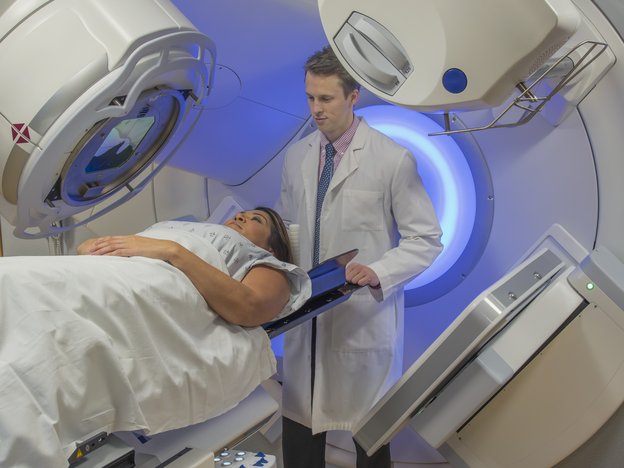 A man in a white coat and blue tie supervises a female patient during an MRI scan. They are in a hospital.