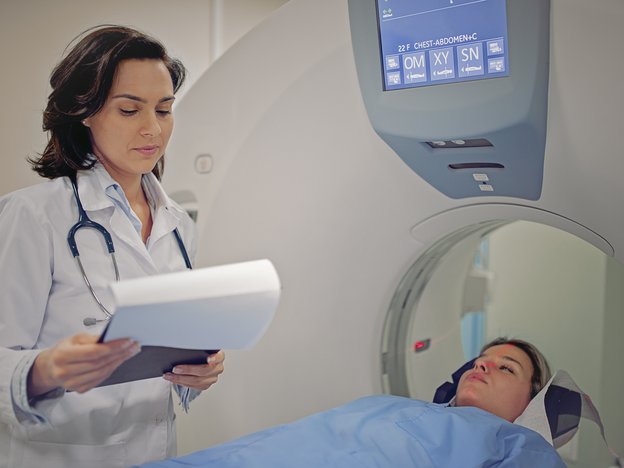 A woman in a white coat holding a clipboard supervises a patient during an MRI scan. They are in a hospital.