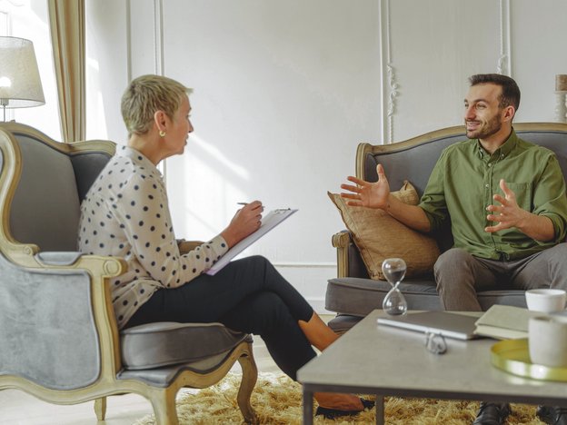 A woman in a white and black spotted shirt and black pants faces a male in a green shirt and grey pants. She is taking notes on a clipboard as they interact.