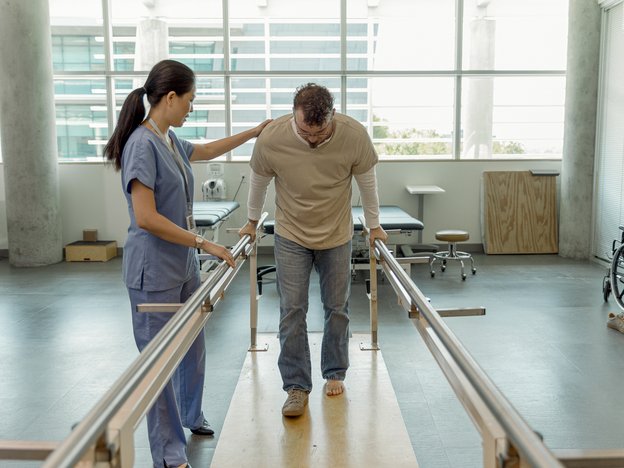 A woman helps a man on crutches walk. The man wears one shoe and is in between two guard rails. The woman wears blue scrubs. They are inside.