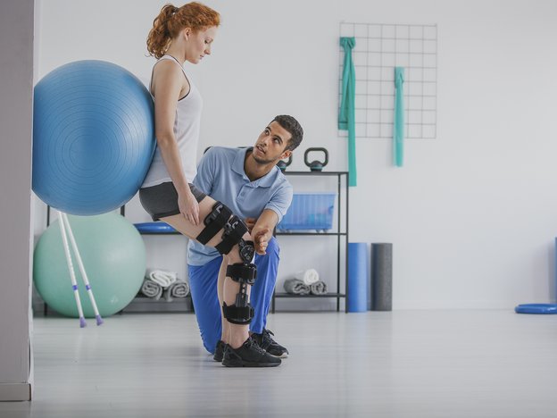 A woman leans on a medical ball against the wall. A man kneels down and places his hand over her knee. She wears a leg brace and the man wears blue uniform