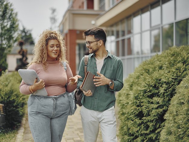 A woman in a pink shirt and blue jeans walks through a school campus with a man in a green shirt and white pants.