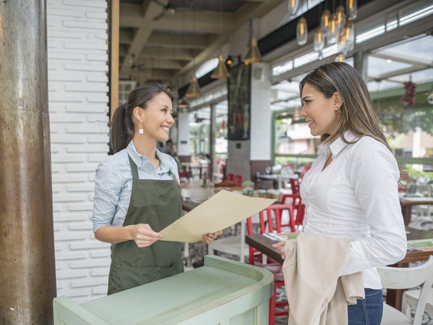 A woman wears an apron and interacts with another woman at a restaurant. The woman with an apron holds a big sheet of paper.