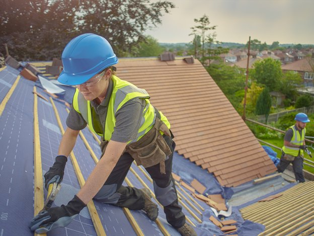 A woman hammers a strip of wood onto a roof of a house. She wears a blue helmet, high visibility vest, tool belt and gloves. There is a male worker behind her.