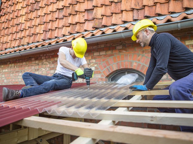 Two men apply part of a roof to a house. They both wear gloves and yellow helmets. One man drills the roofing material onto wood.