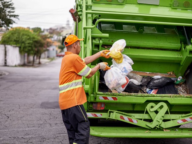 A man in an orange and yellow shirt with an orange hat loads rubbish into a rubbish and recycling truck on a road.