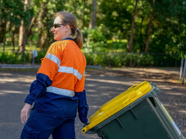 A woman in an orange shirt drags a green rubbish bin with a yellow lid outdoors.