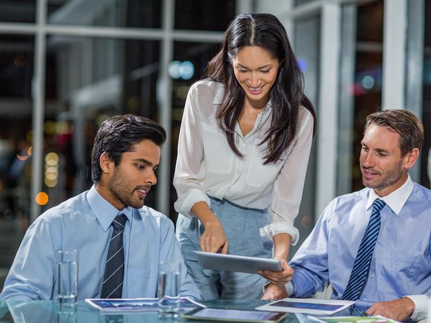 Three business professionals look at a tablet. They are wearing formal work clothes in a corporate office.