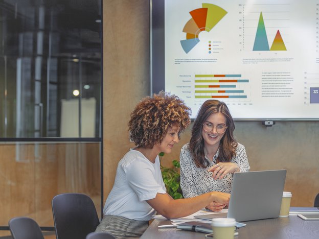 Two sales professionals view laptop in a modern office. Graphs are displayed on a projector behind them.