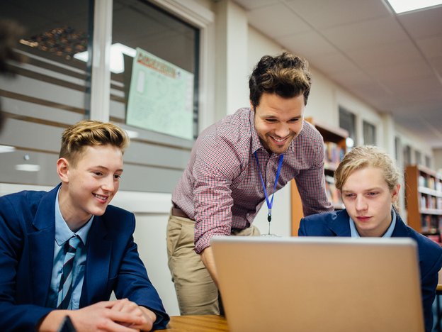 A male student looks at an open laptop on a classroom table. A male teacher leans over and helps him. Another male student is sat next to the teacher.