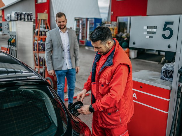 A man closes a fuel cap on a car at a petrol station. Ge wears red overalls and a red vest. There is a man behind him, he wears a blazer.