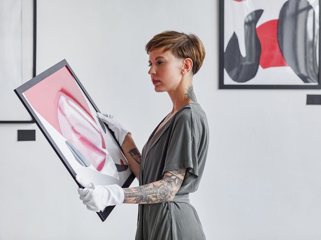A woman is holding a piece of art wearing white gloves. She stands in a gallery.