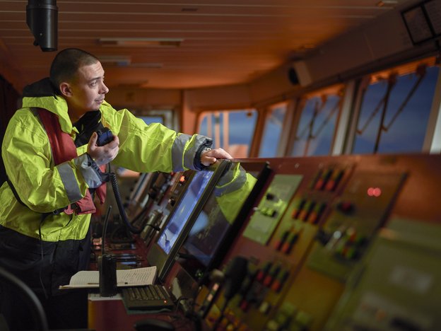 A man leans over a navigation system in a ships control room. He wears a yellow high visibility jacket and holds a walkie talkie attached to the system.