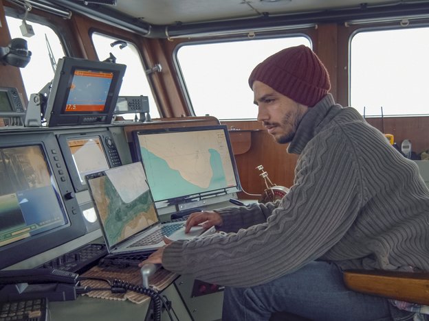 A ship master uses a laptop as he observes three monitors on a ship. He wears a grey jersey and red beanie.