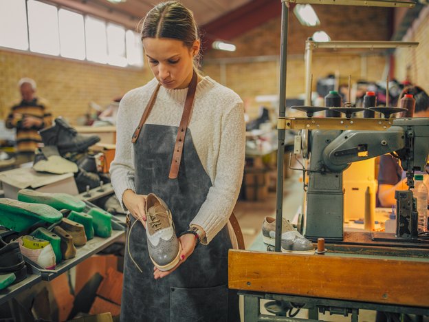 A woman holds a shoe. She wears an apron and stands inside a workshop. She is surrounded by shoes and machinery.